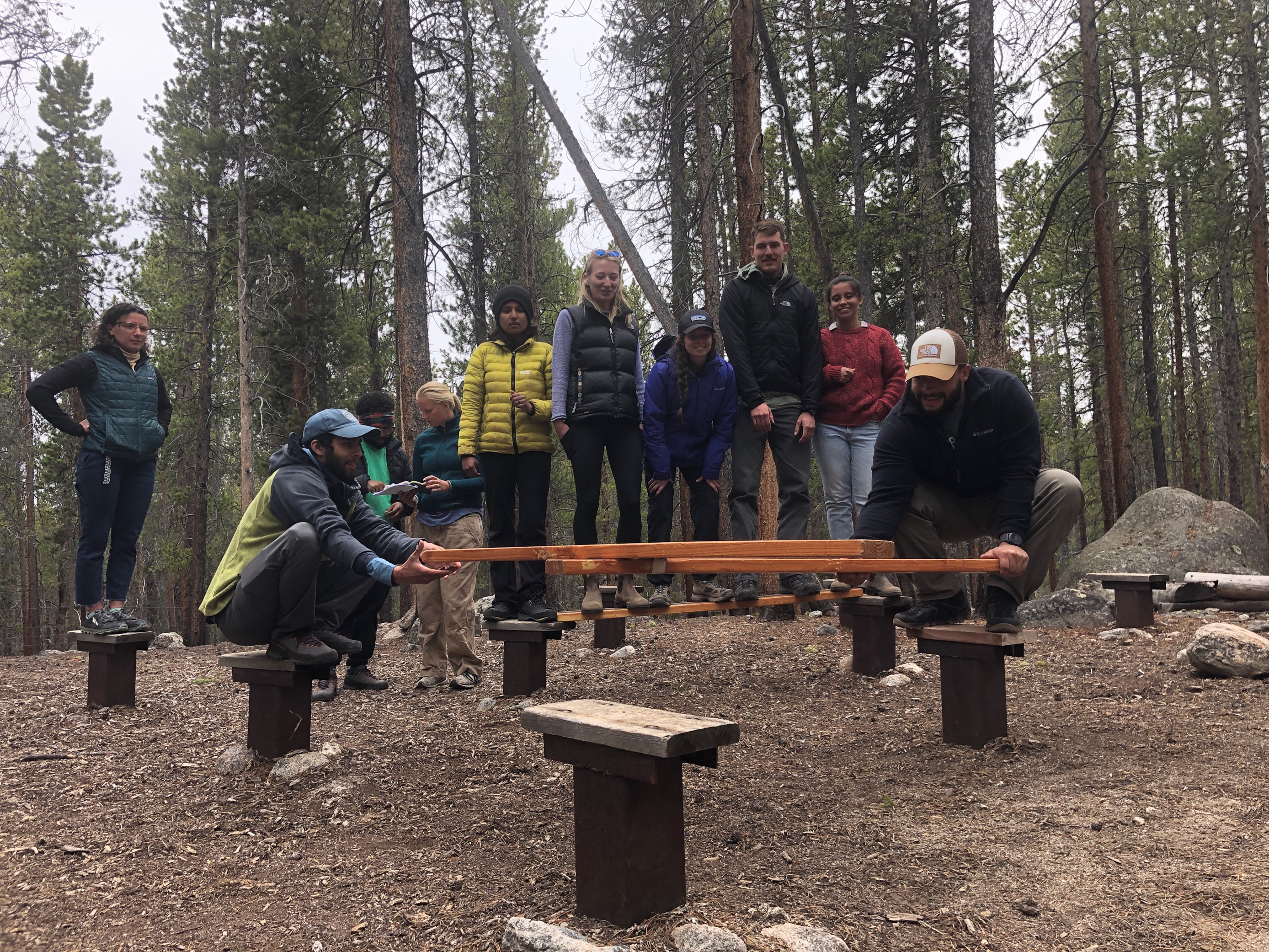A group of people are standing on logs and other wooden platforms in the forest as part of a low-ropes initiative. Most of the group looks on while a pair of people move two boards in an effort to create a bridge for the others to pass. They are all wearing pants and jackets.